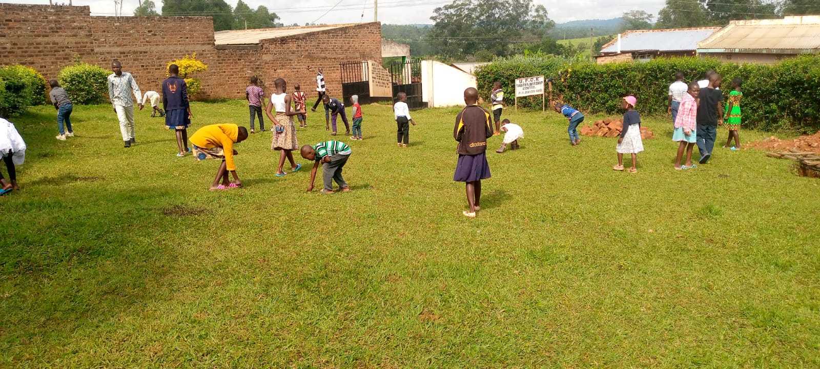 Children and adults cleaning a grassy compound outdoors.